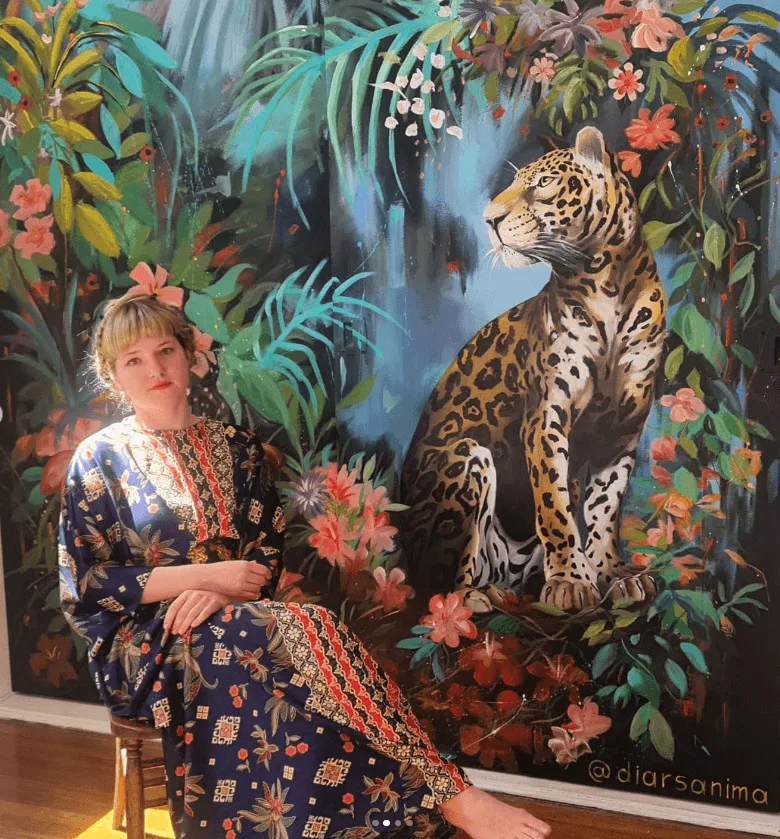 DiArsanima seated in front of her painting of a jaguar in a tropical jungle